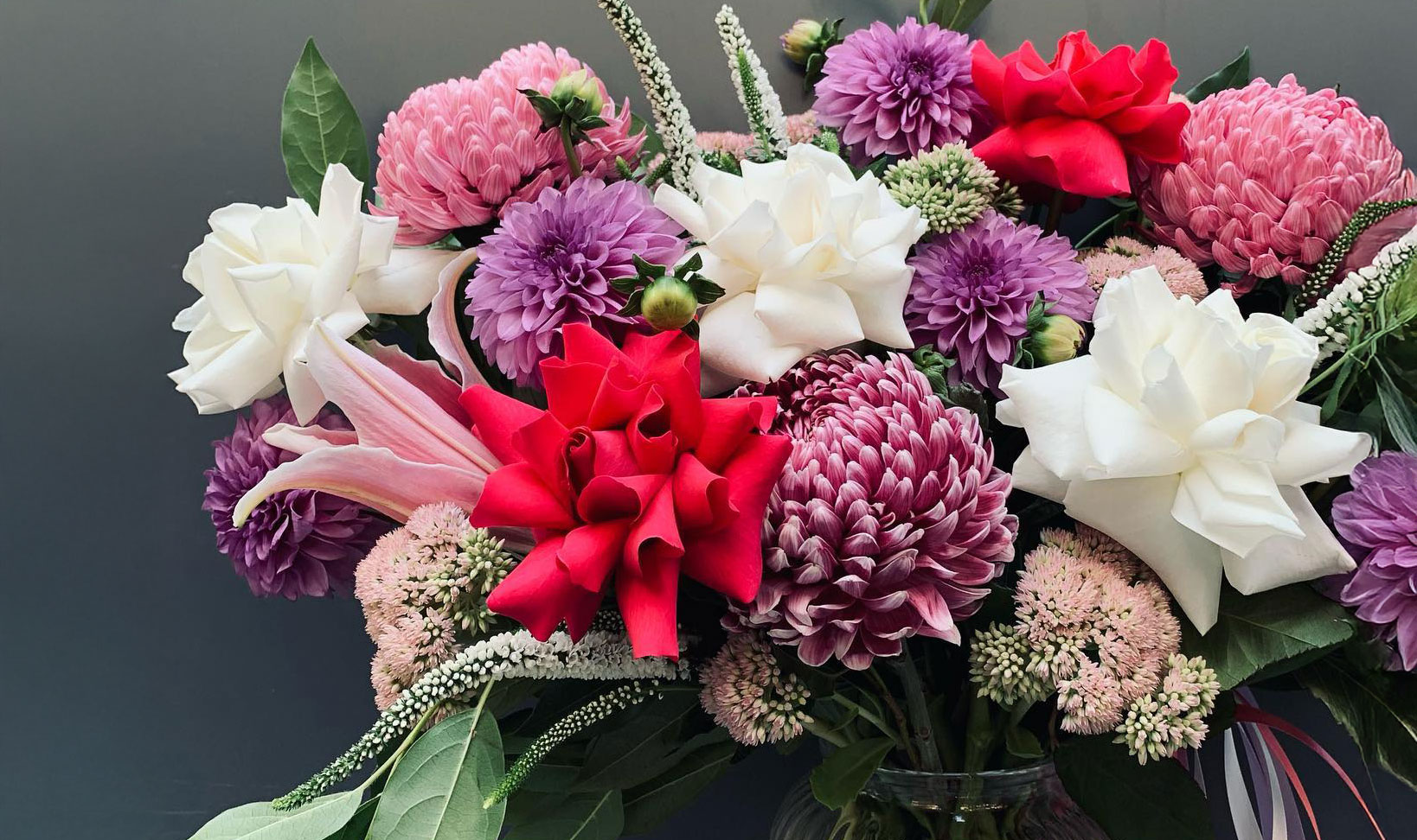 A bouquet of fresh red, pink, white, and purple flowers with green leaves stands vibrant against a dark background, perfect for celebrations.